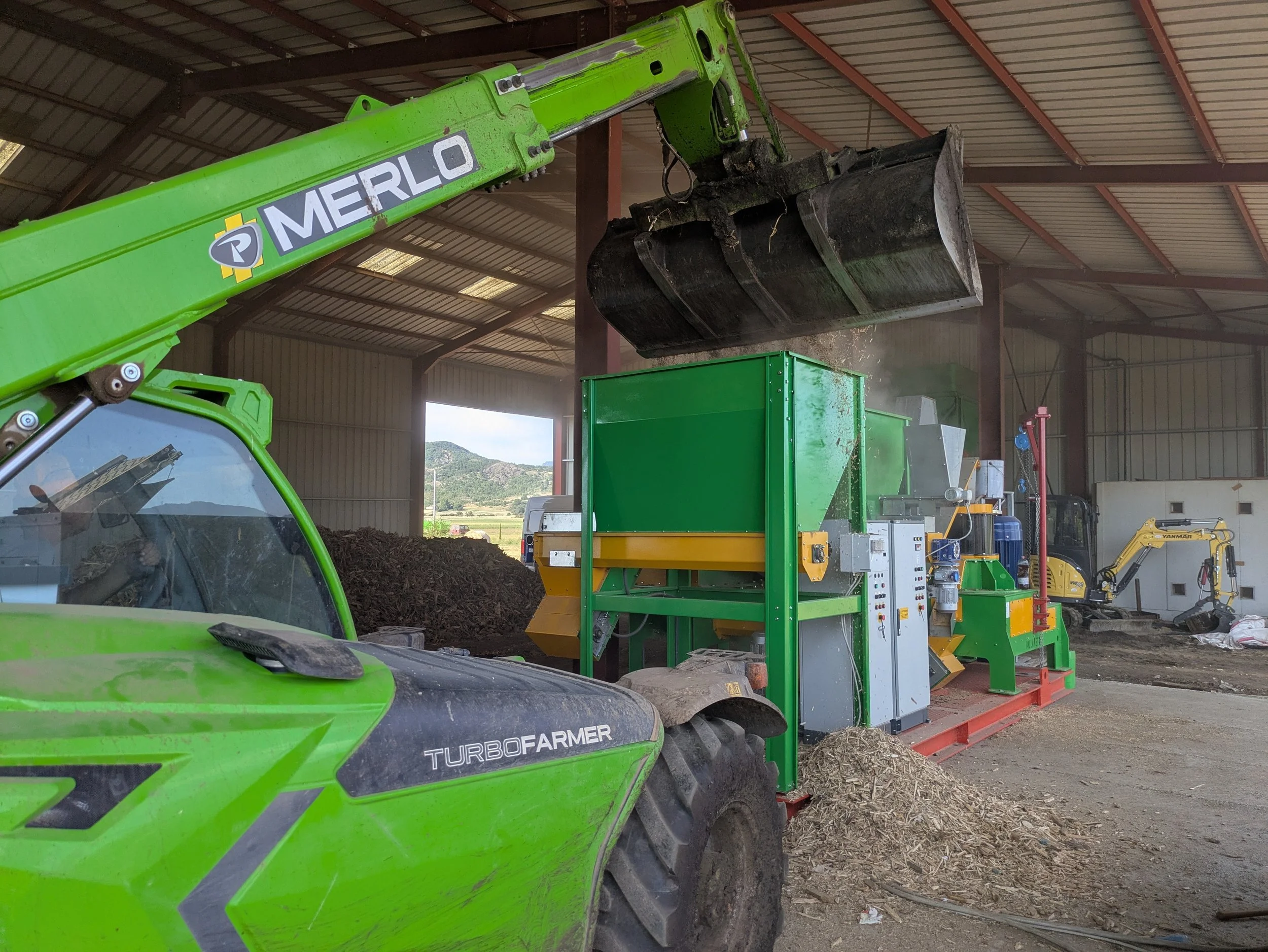 Une machine agricole en intérieur, avec un bras mécanique vert portant une grande cuve, une autre machine à côté, toutes deux dans un environnement industriel, avec un sol en terre et des montagnes en arrière-plan.
