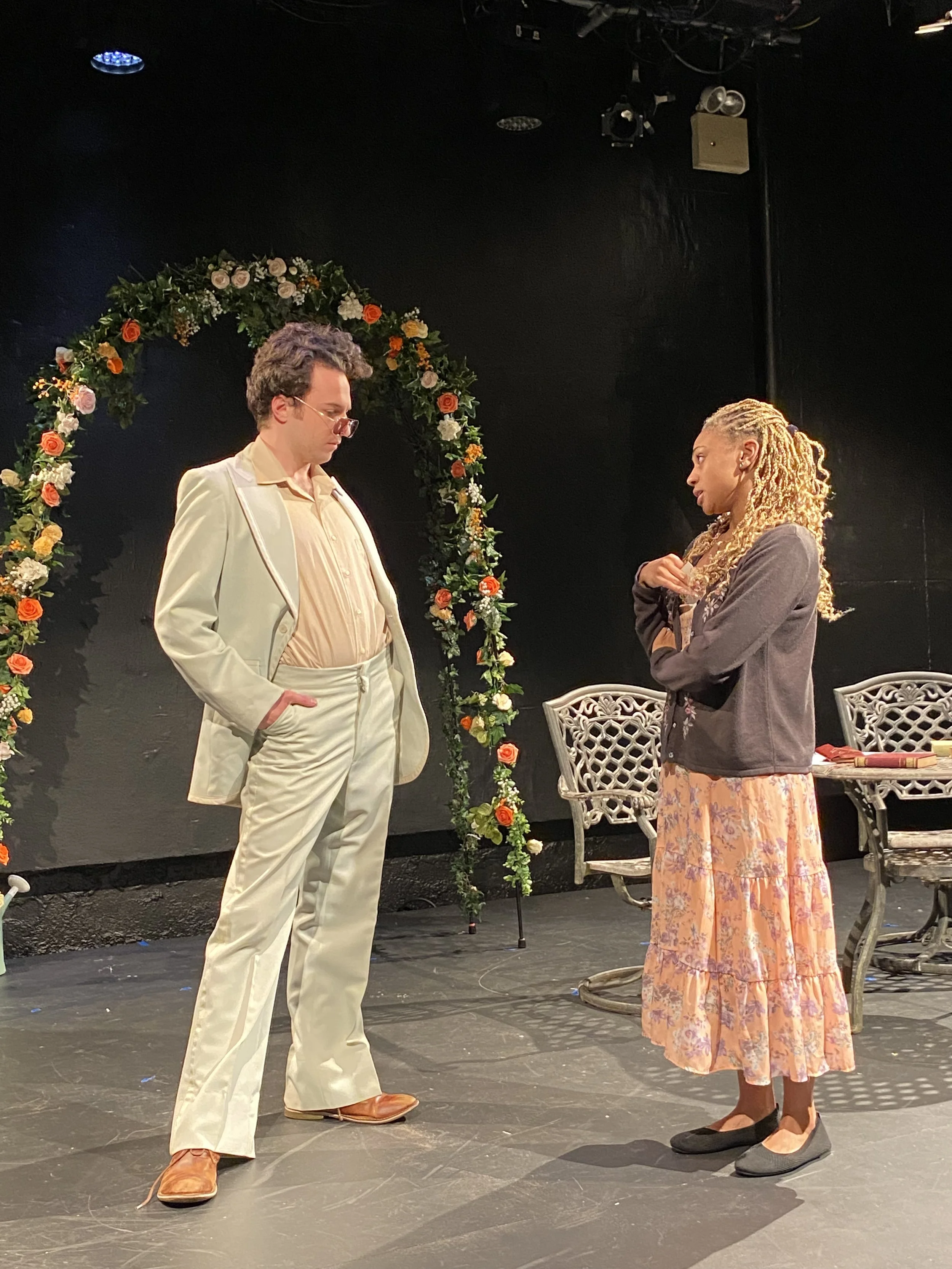 Two actors perform a scene on stage, one man in a cream suit with sunglasses and a woman in a floral skirt and gray cardigan. They stand near a floral arch and a bench, with stage lights overhead.