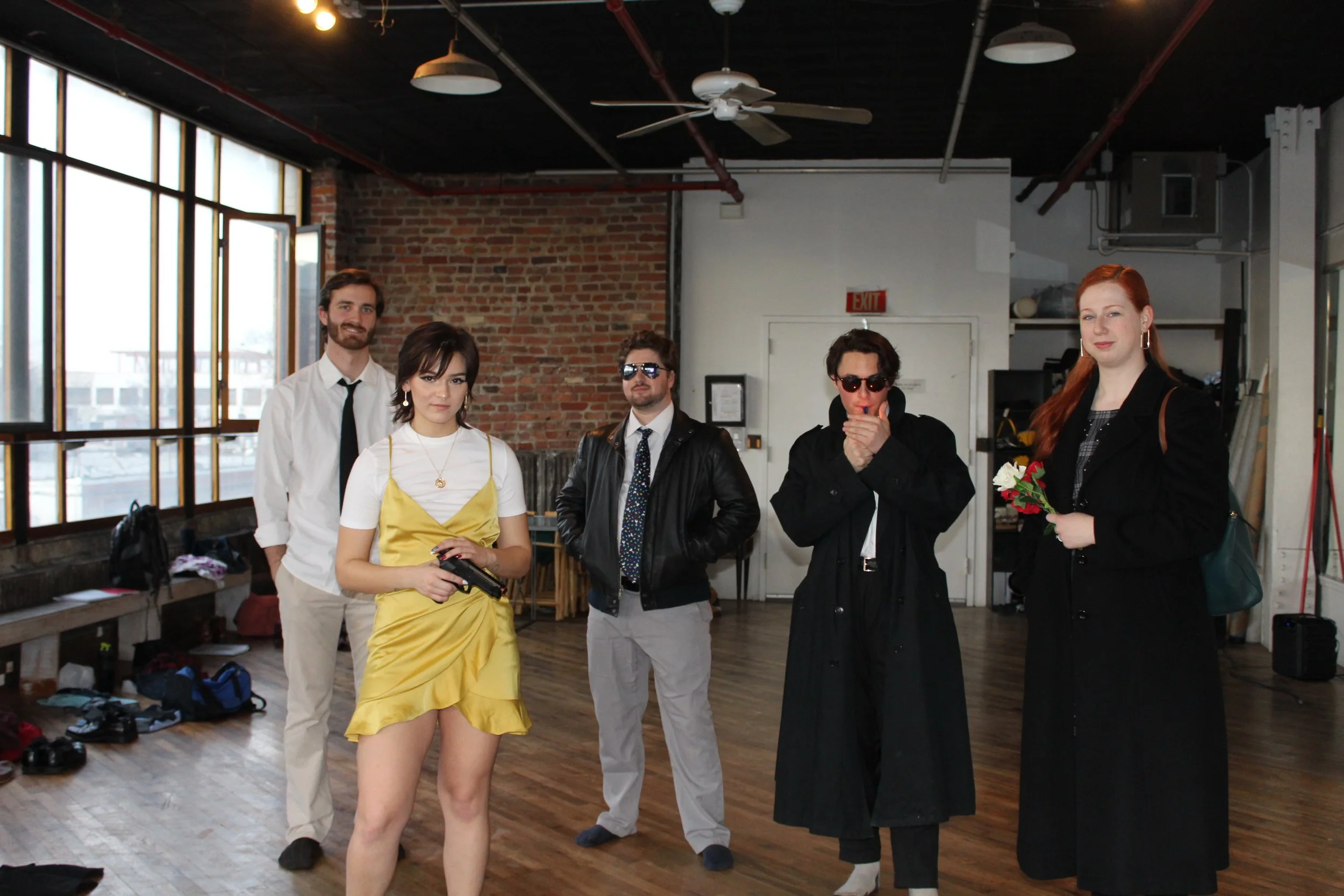 Five people standing in an indoor space with a brick wall and windows, some holding items like a gun and flowers, dressed in a mix of casual and stylish clothing.