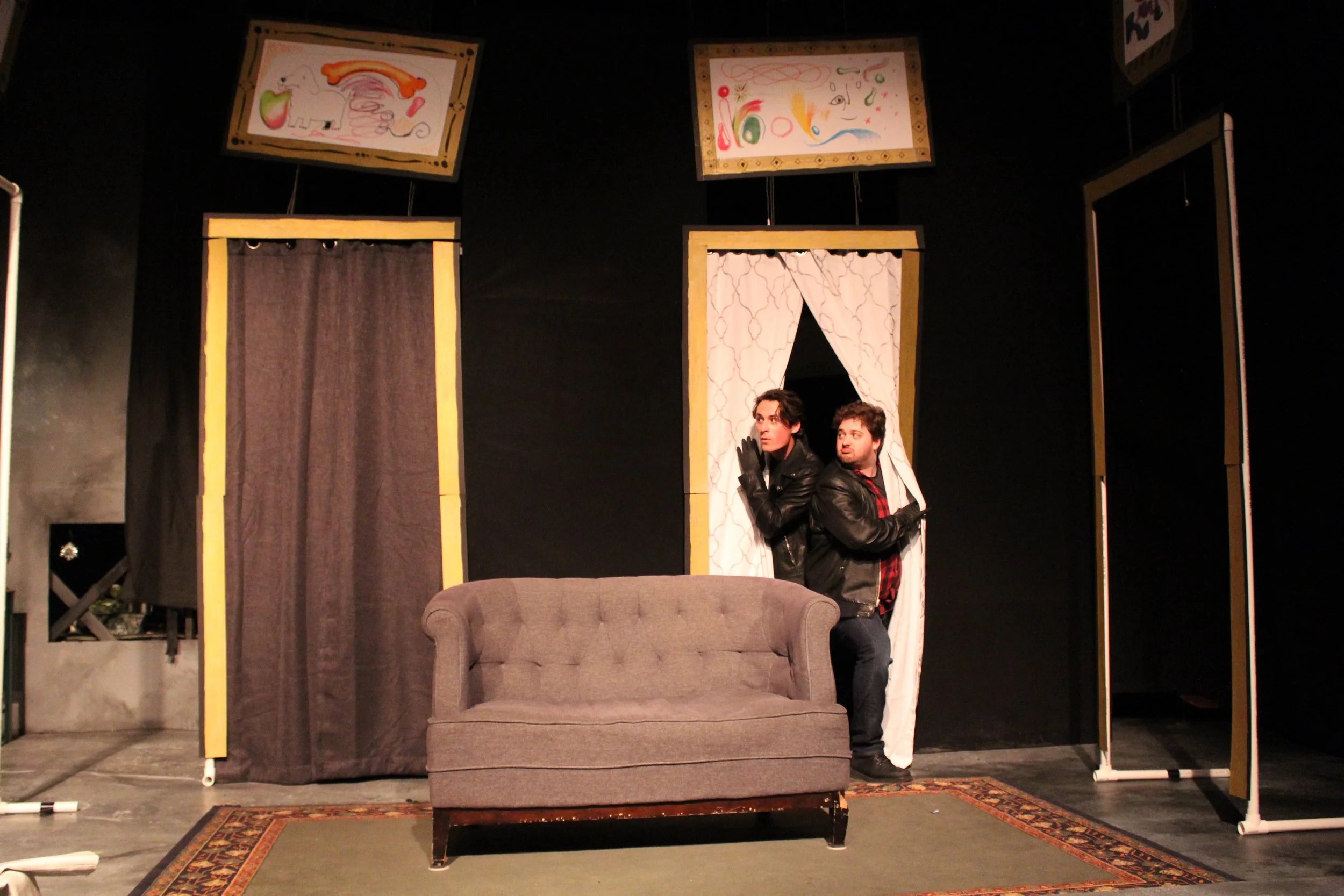 Two men in leather jackets stand behind a gray couch on a stage with a black background and colorful abstract art above. They are peeking out from behind curtains, with one man looking to the side and the other looking straight ahead.