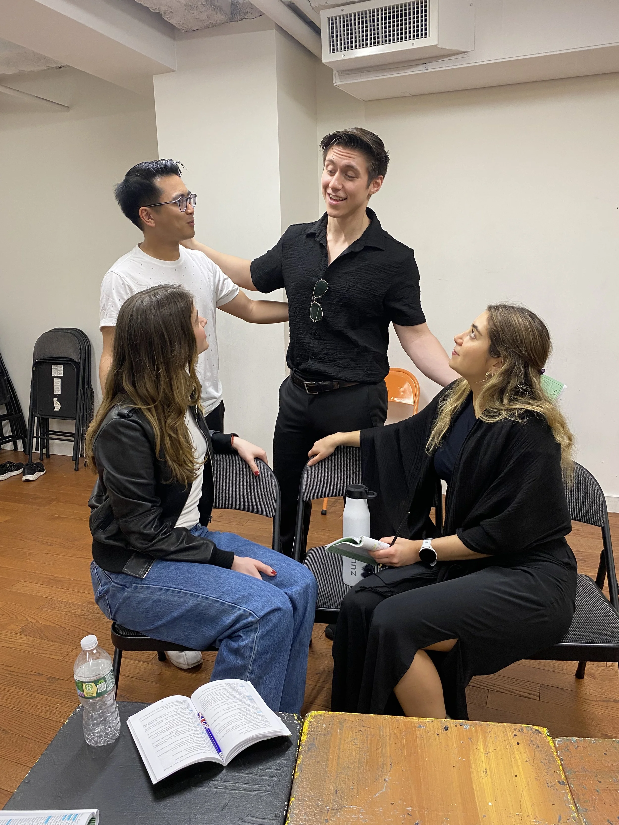 A group of five people engaged in a conversation in a room, with one person standing and four people sitting; two women and one man are seated while one man stands with arms outstretched, smiling.