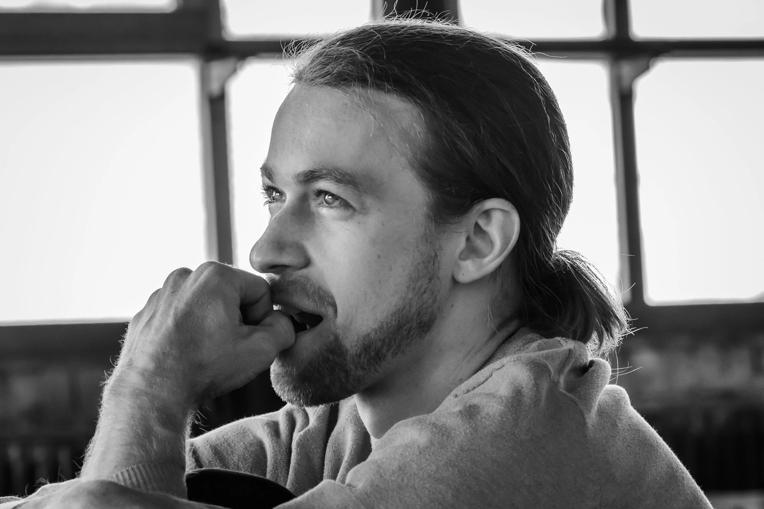 A young man with long hair and a beard, resting his chin on his hand, looking thoughtfully out a window in a black-and-white photo.