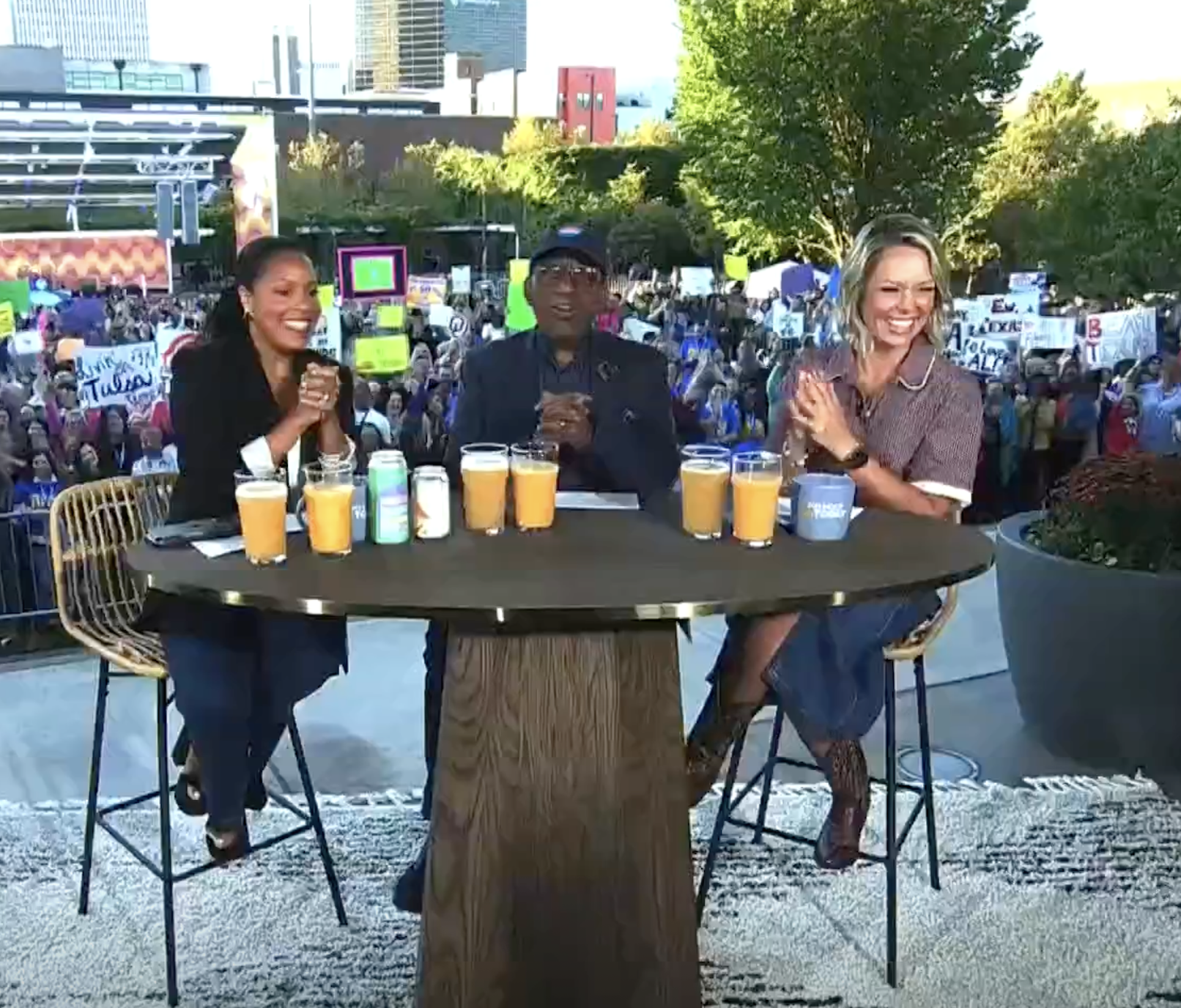 Three people sitting at a table with drinks, on a stage with a crowd of protesters and city buildings in the background.