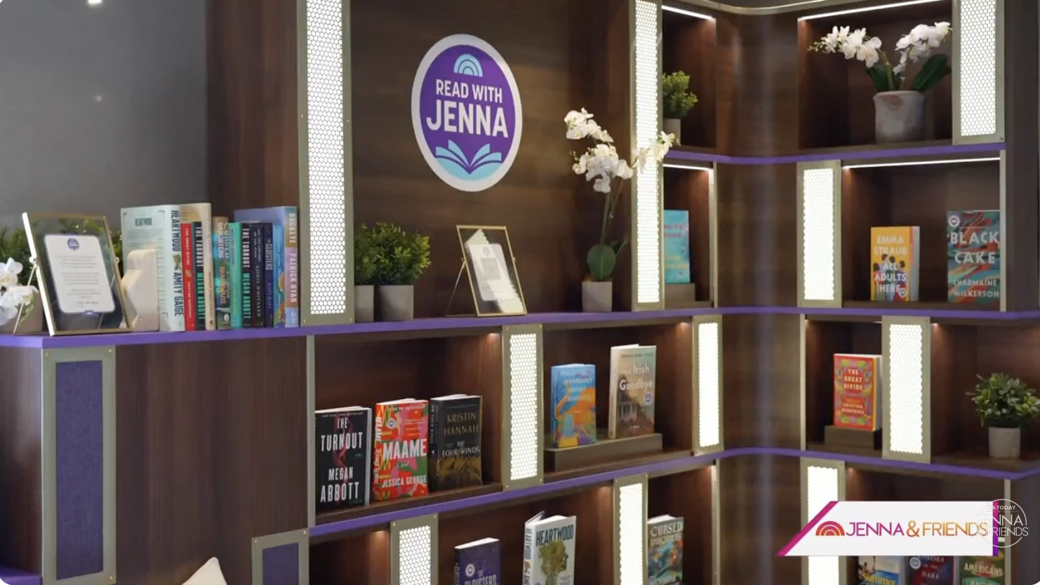 A bookshelf with books, plants, and framed pictures in a wooden room with purple accents and a sign that reads 'Read with Jenna.'