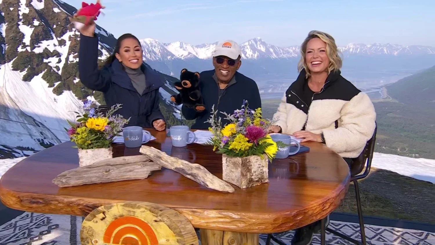 Four smiling people sitting at a table outdoors with mountain scenery, wearing winter jackets, and flower arrangements, celebrating on a TV show set.