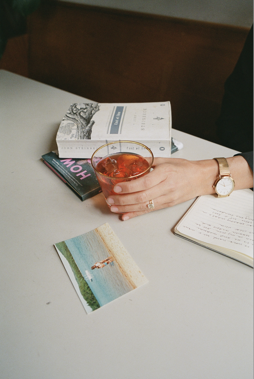 A person resting their hand on a table, holding a glass of iced tea. The table has a book, a magazine, a postcard, an open notebook with handwriting, and a wristwatch. The person's hand has a ring, and they are wearing a gold watch.