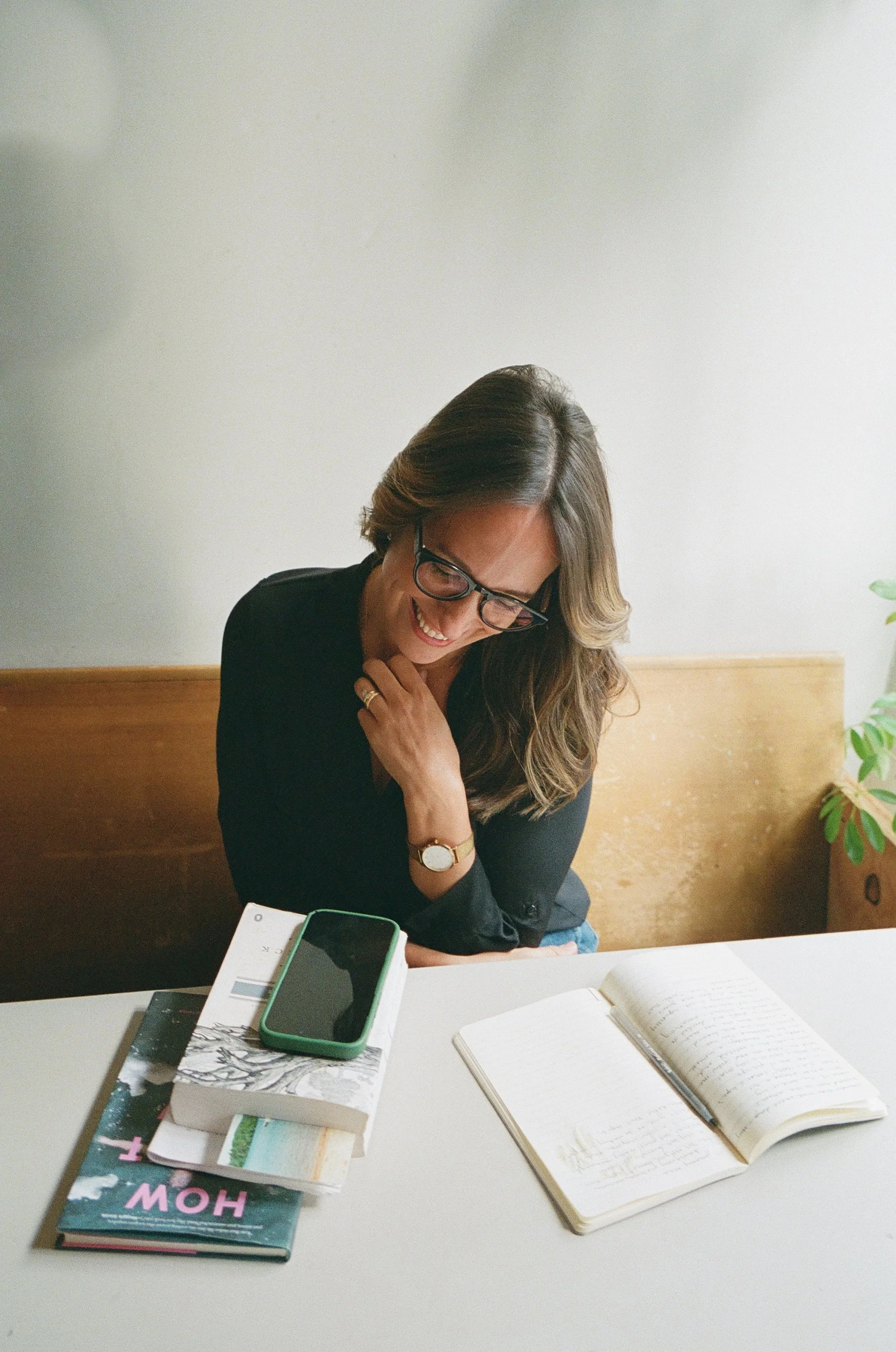 A woman with glasses smiling, sitting at a table with a notebook, a smartphone, and several books.