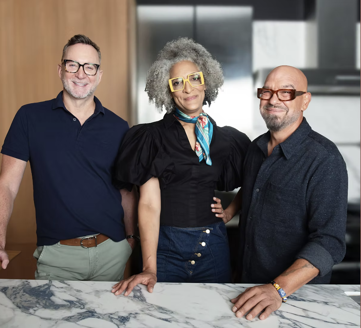 Three adults standing together in a kitchen, smiling at the camera. The man on the left has glasses, short brown hair, and a beard, wearing a navy blue polo shirt and khaki pants. The woman in the middle has curly gray hair, yellow glasses, and is wearing a black top with puffy sleeves and a colorful scarf. The man on the right has a bald head, glasses, a gray beard, and is wearing a dark button-up shirt with the sleeves rolled up.