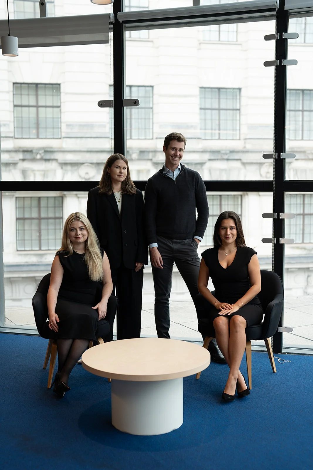 Five business professionals in an office with large windows, two seated women in black dresses, one standing woman in a black blazer, and two standing men, one smiling.