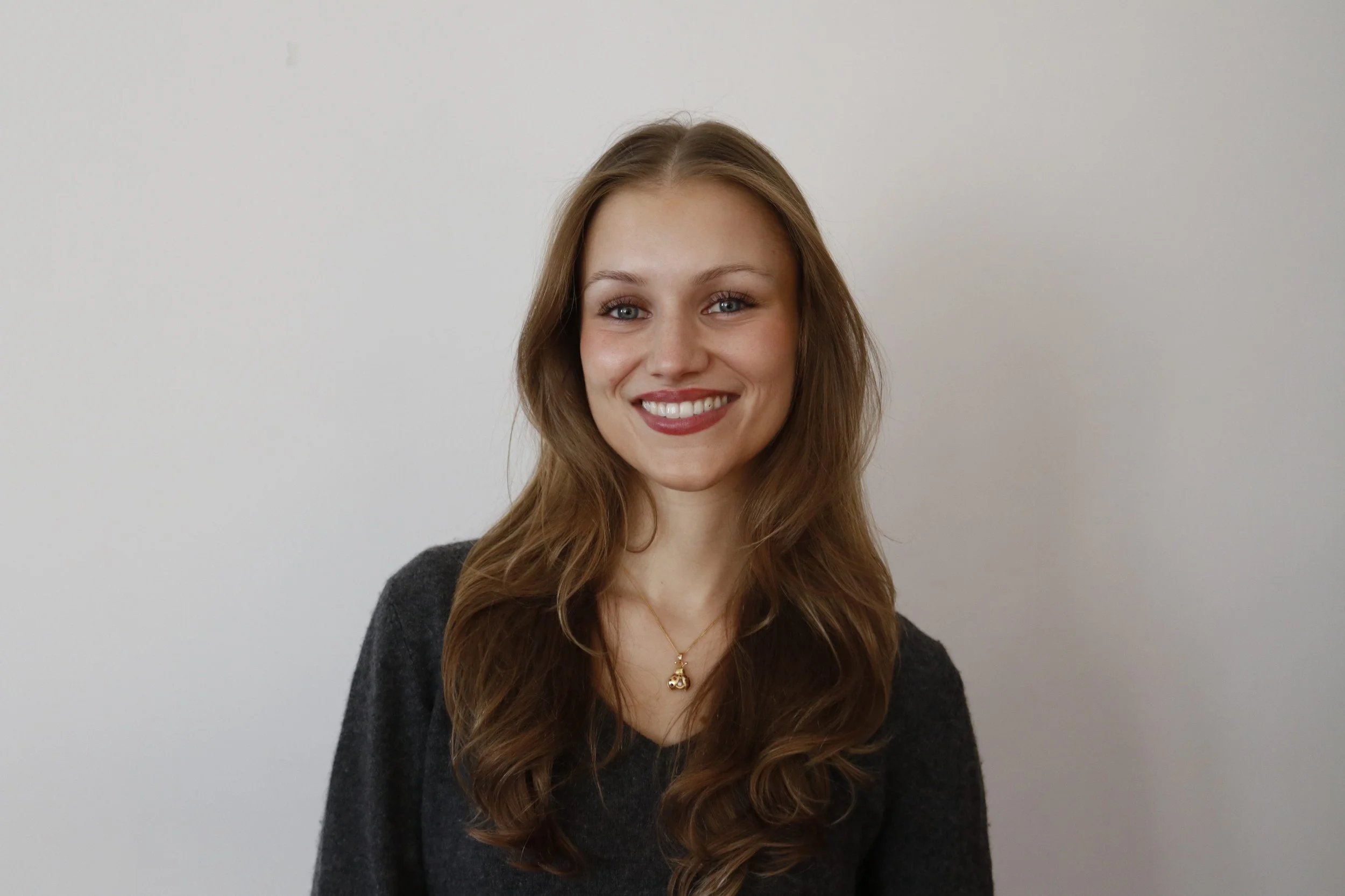 A young woman with long wavy brown hair, blue eyes, and wearing a dark gray sweater and gold necklace, smiling against a plain white wall background.