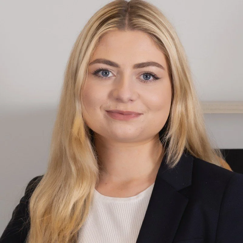 A blonde woman with long hair, blue eyes, wearing a black blazer and white top, smiling slightly, in an indoor setting.