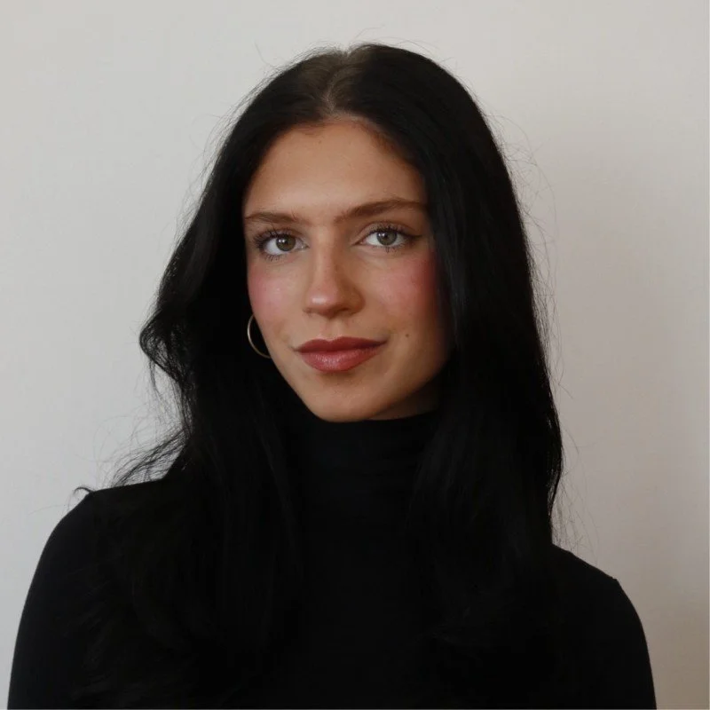 A woman with long black hair wearing a black turtleneck and hoop earrings, standing against a plain white background.