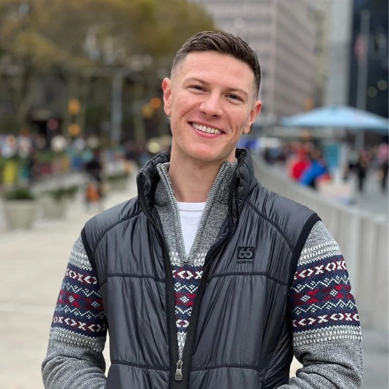 Young man smiling outdoors in an urban area, wearing a patterned sweater, a gray zip-up jacket, and a black vest.