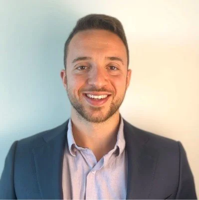 A young man with dark hair and a beard wearing a navy blue suit and light-colored shirt, smiling against a light background.