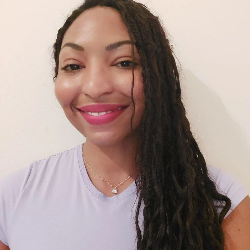 Close-up of a woman smiling, wearing a lavender top and a necklace with a pendant, with long curly hair over her shoulder.