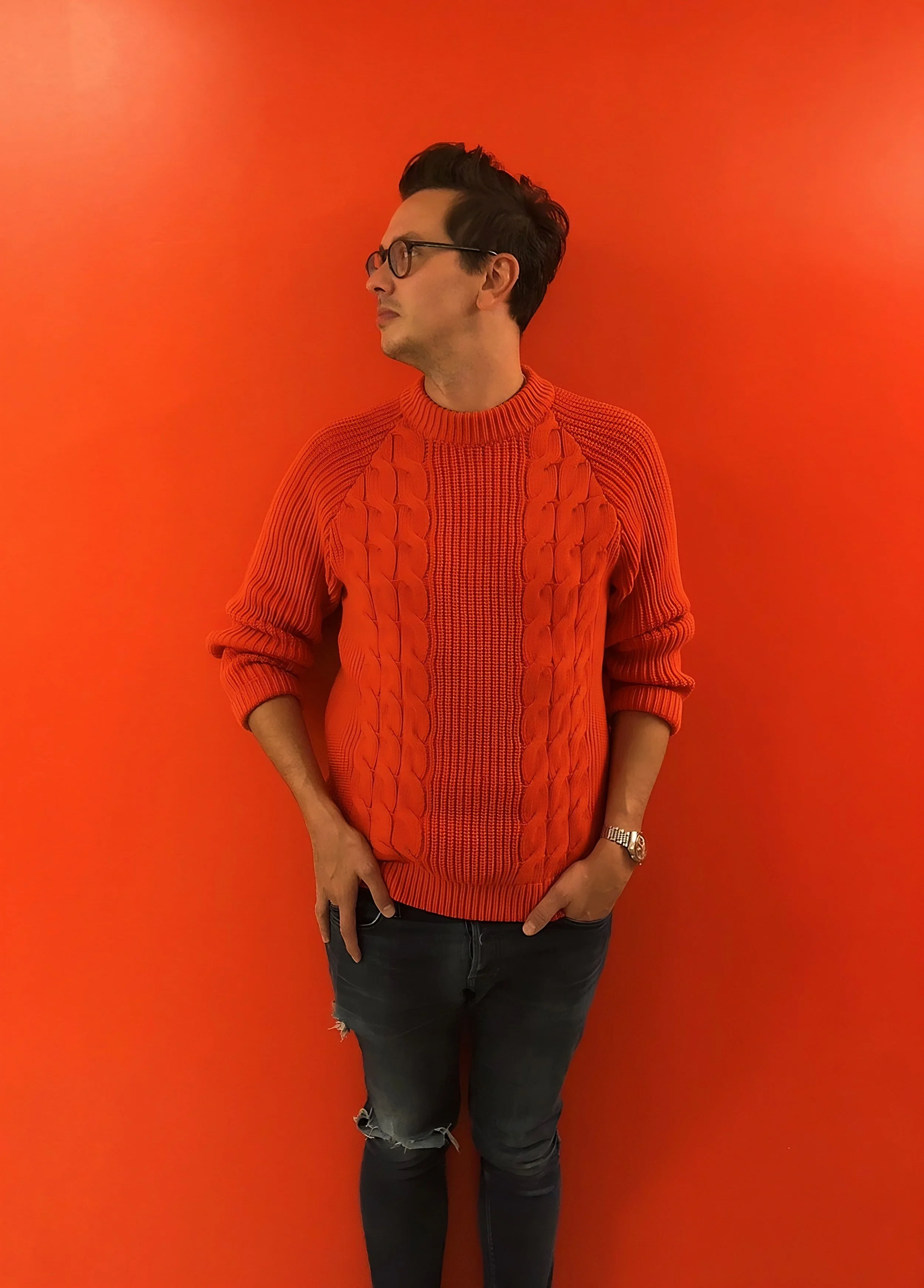 A man with dark hair, glasses, and light skin wearing a red sweater with a cable-knit pattern and distressed black jeans, standing against an orange wall.