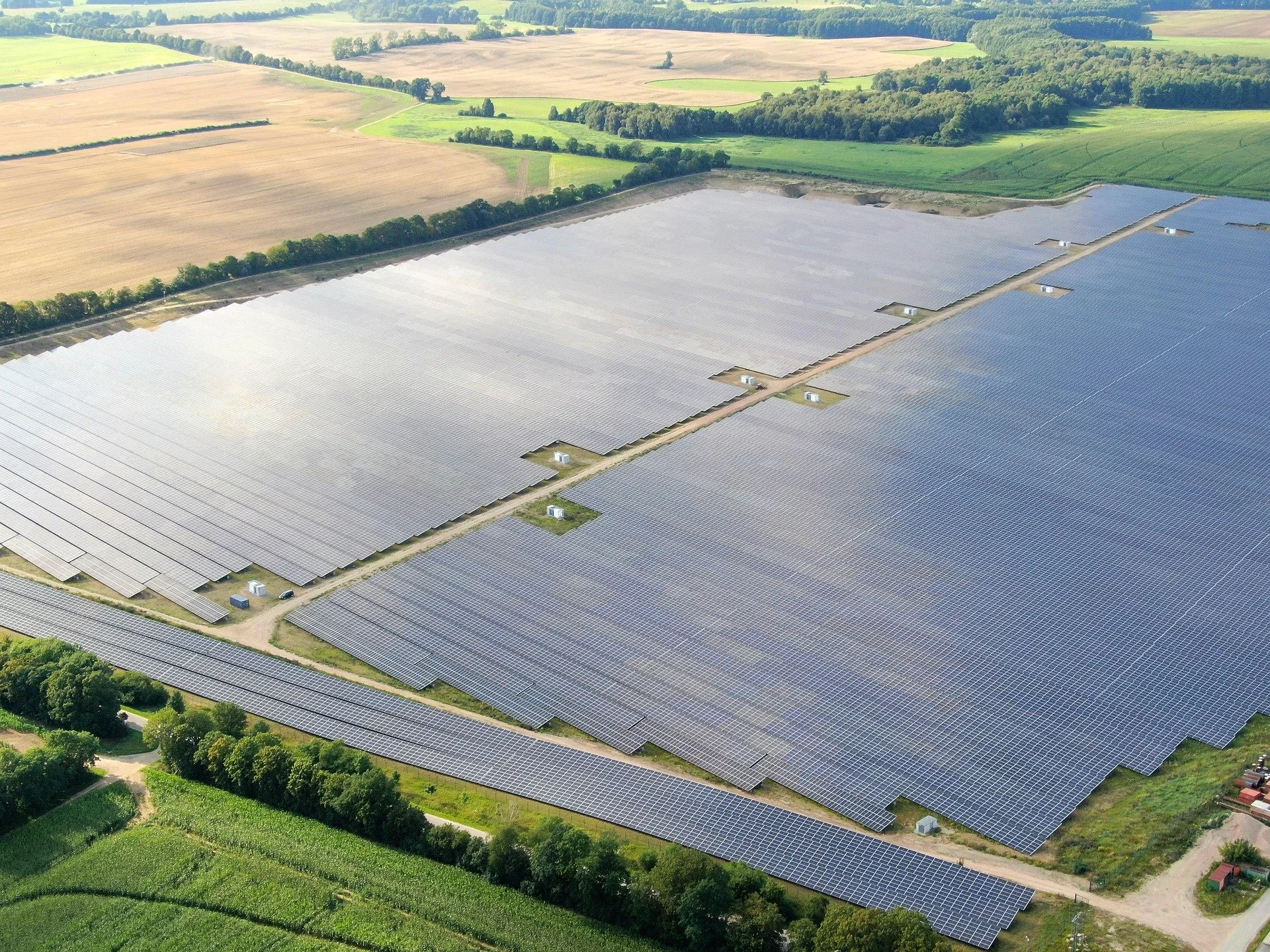 Vogelperspektive auf ein großes Solarpark mit vielen Solarmodulen, umgeben von landwirtschaftlichen Feldern und Wäldern.