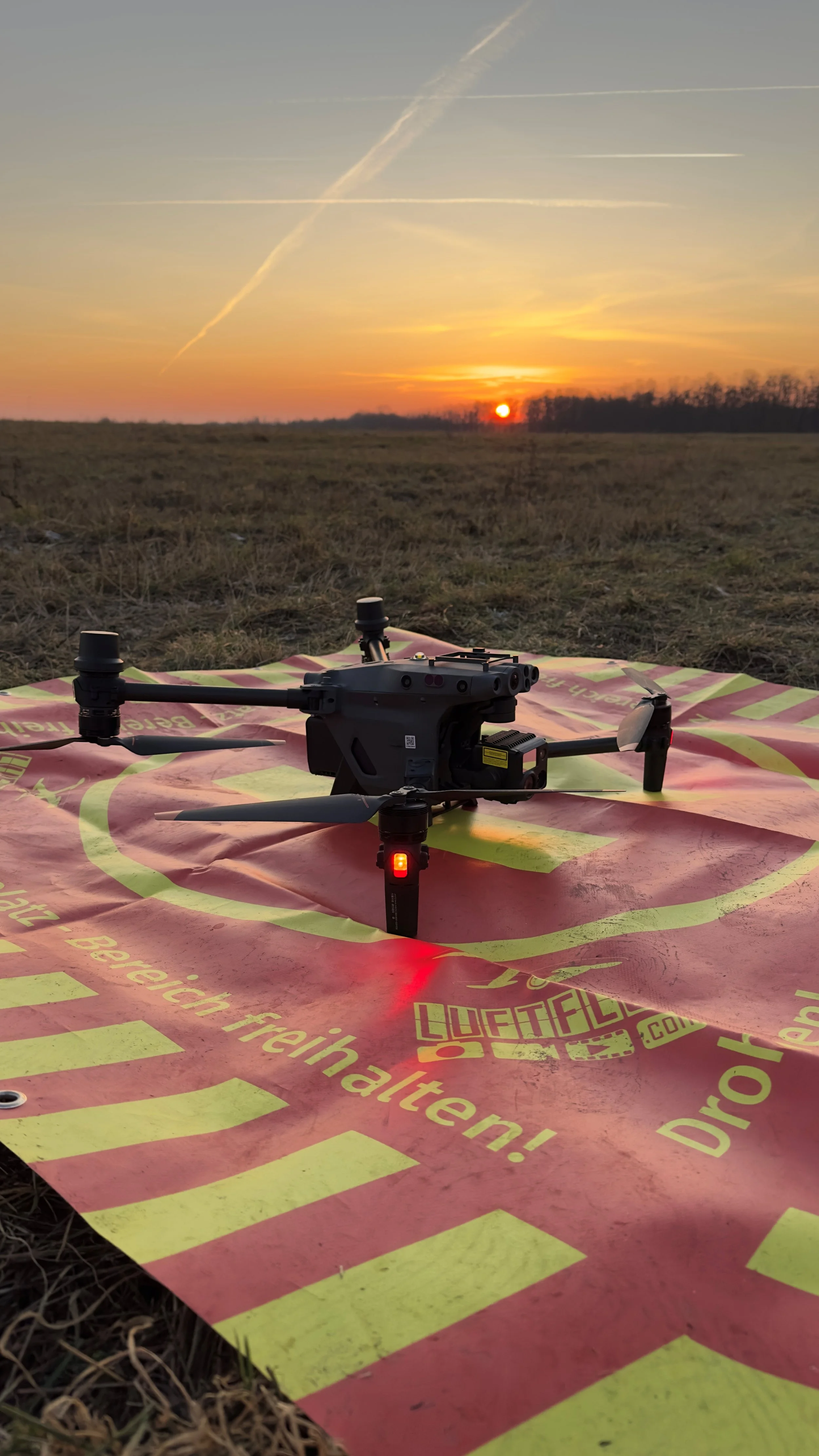 Drohne auf einer rot-gelben Bodenplane bei Sonnenuntergang mit Himmel und Wolken im Hintergrund.