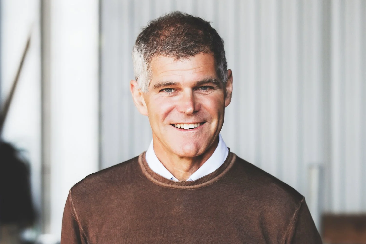 A smiling middle-aged man with short, gray hair, wearing a brown sweater over a white collared shirt, standing in front of a light-colored, corrugated metal background.