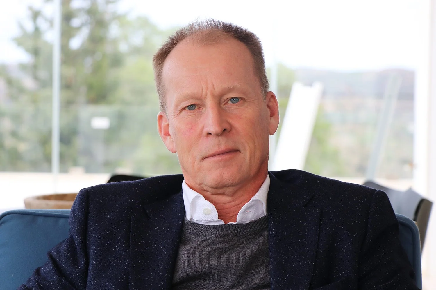 Portrait of a middle-aged man with short hair, wearing a white shirt, dark sweater, and blazer, sitting indoors with a blurred background of windows and trees.