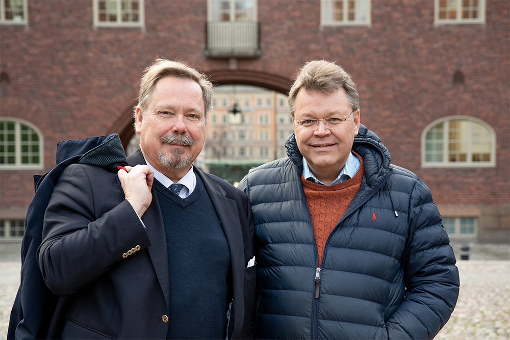 Two men standing outdoors in front of a brick building, smiling at the camera. One is wearing a dark suit and carrying a shoulder bag, the other a navy puffer jacket and layered sweater.