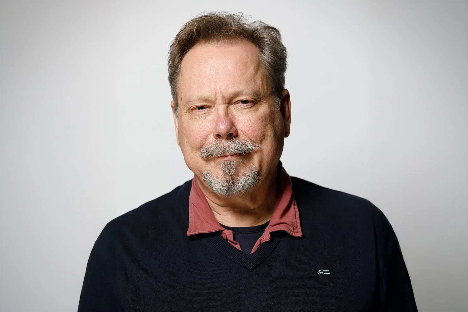 Portrait of an older man with gray hair, a mustache, and a goatee, wearing a dark sweater over a red collared shirt, against a plain light background.