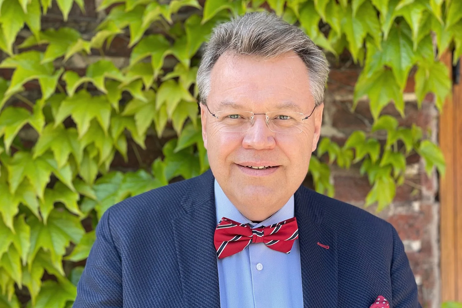 A man wearing glasses, a navy blazer, a light blue shirt, and a red bow tie with black and white stripes, smiling outdoors in front of green ivy on a brick wall.