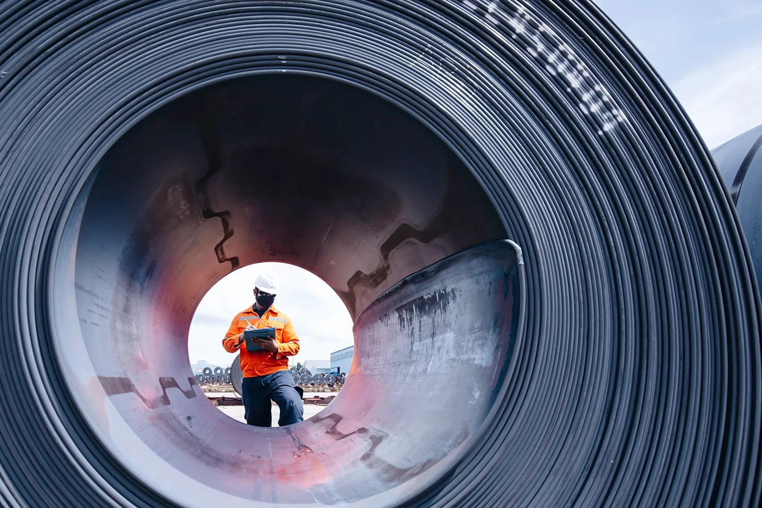 A worker in orange safety gear and a white hard hat inspects large metal coils outdoors, using a tablet device.