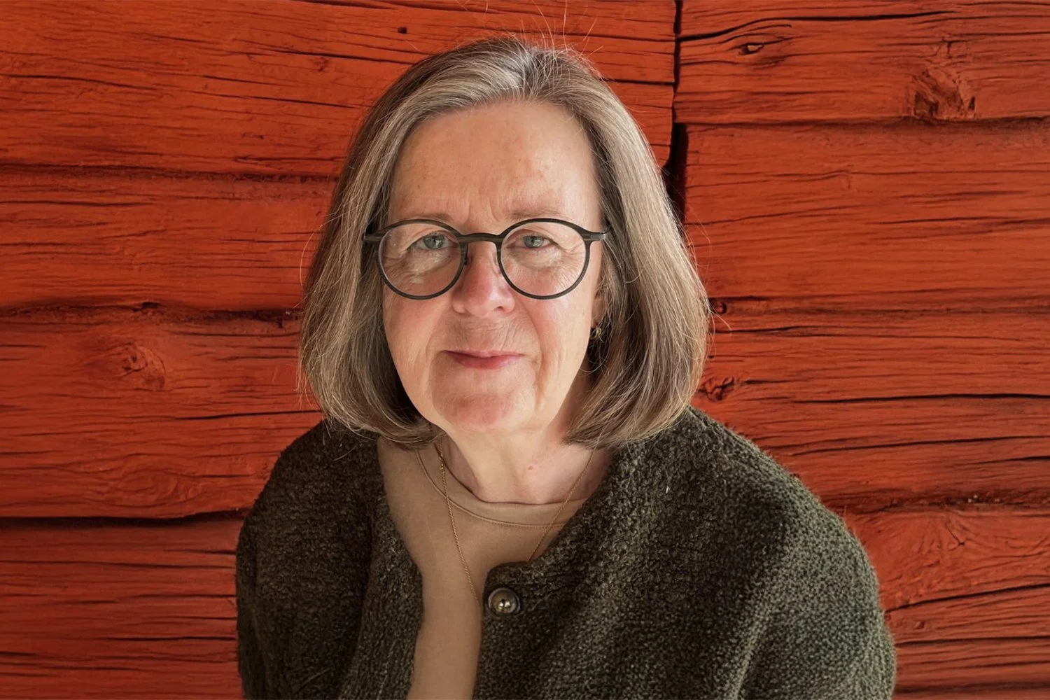 A woman with shoulder-length gray hair and glasses, wearing a brown textured sweater, sitting in front of a red wooden wall.