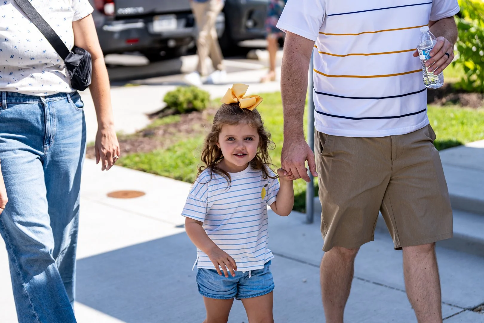 A young girl with a large yellow bow in her hair holding hands with an adult man while walking on a sidewalk on a sunny day. The girl is wearing a striped shirt and shorts. The man is wearing a striped shirt and shorts and holding a water bottle.