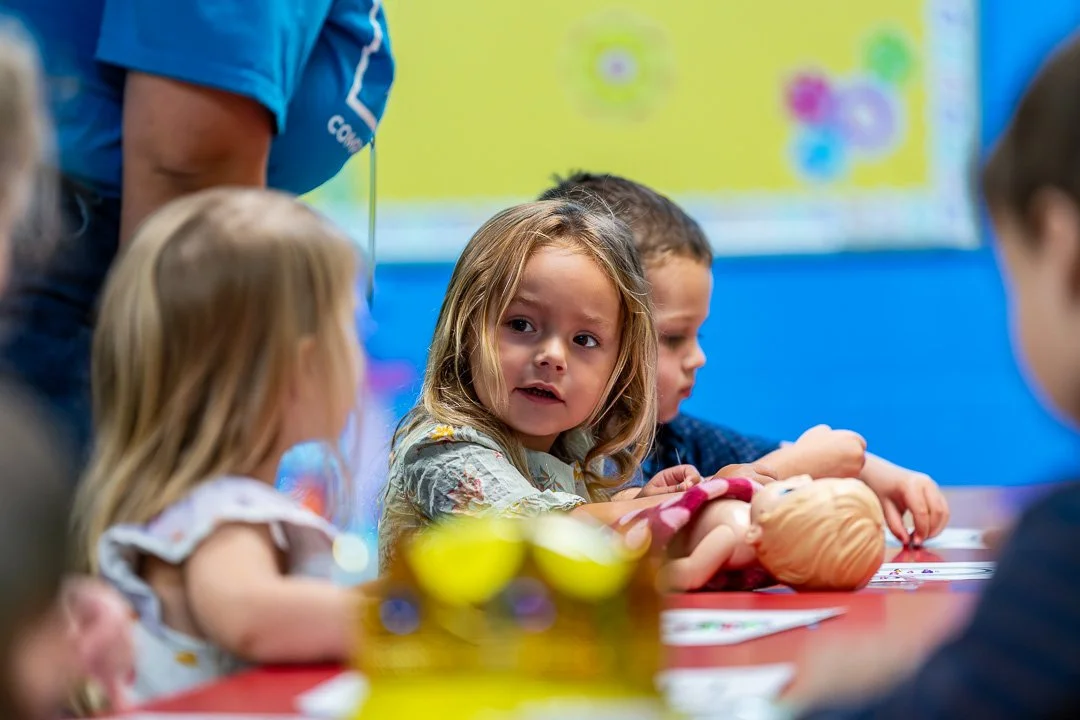 Children gathered around a table in a classroom, with a girl in a floral shirt looking towards the camera, playing with a doll.
