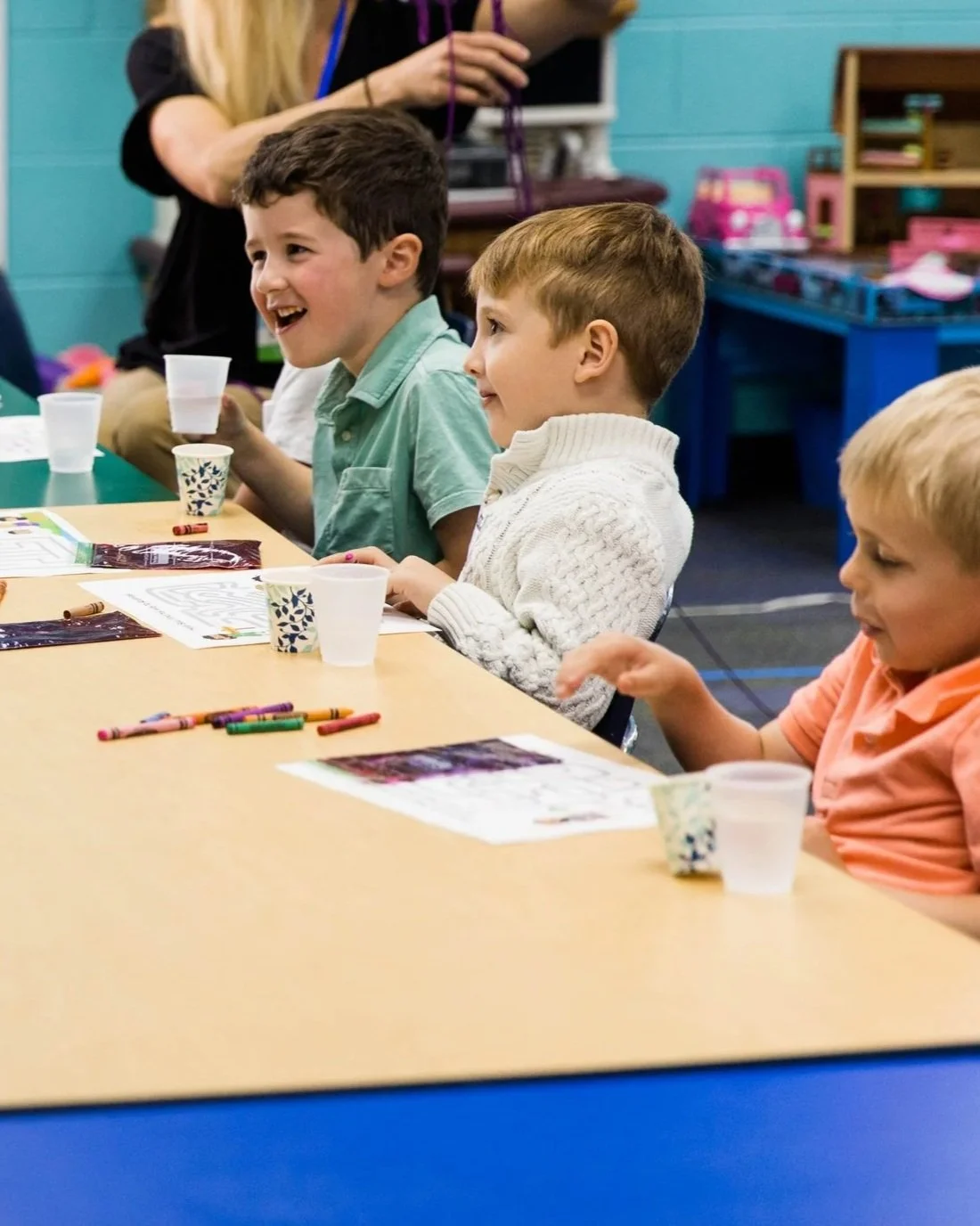 Children sitting at a table in a classroom, smiling and engaging in an activity with crayons and papers, with cups on the table.