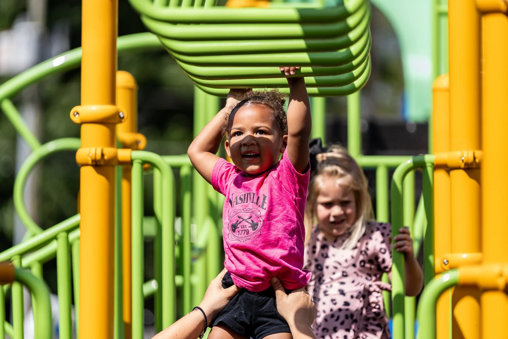 Two young girls playing on a colorful playground with yellow and green bars. The girl in the foreground, with curly hair and a pink shirt, is hanging from the bars, supported by an adult's hands. The girl in the background, with blonde hair and a polka-dotted shirt, is standing near the structure.