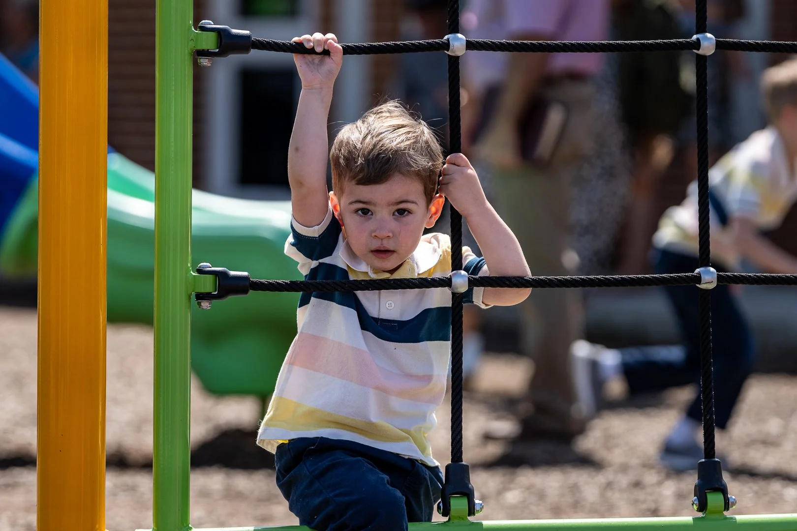 Young boy with brown hair wearing a striped polo shirt climbing on a black rope ladder at a playground.