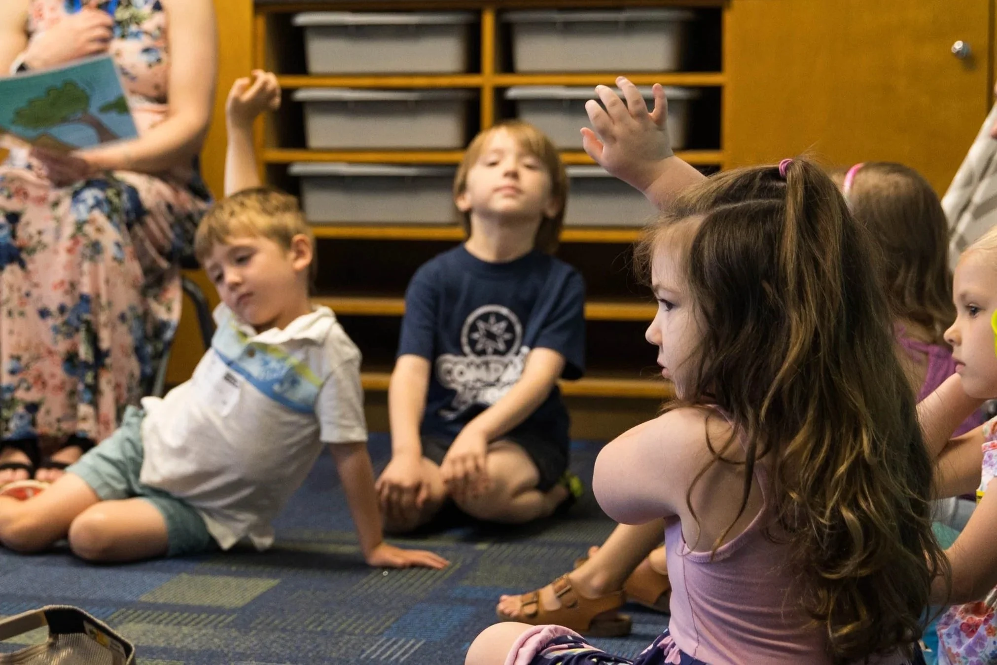 Children sitting on a classroom carpet, with one girl raising her hand, and a woman reading a book in the background.