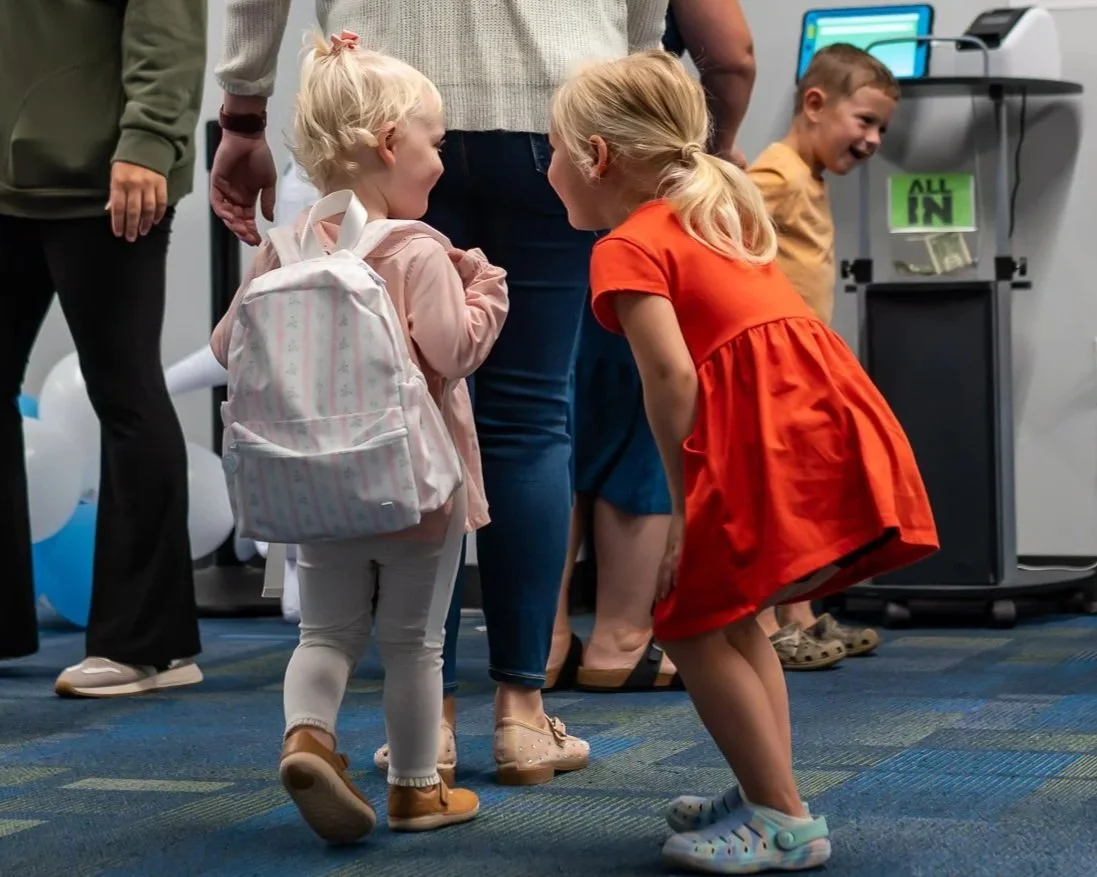 Children laughing and playing together indoors, with some wearing backpacks, in a casual setting.