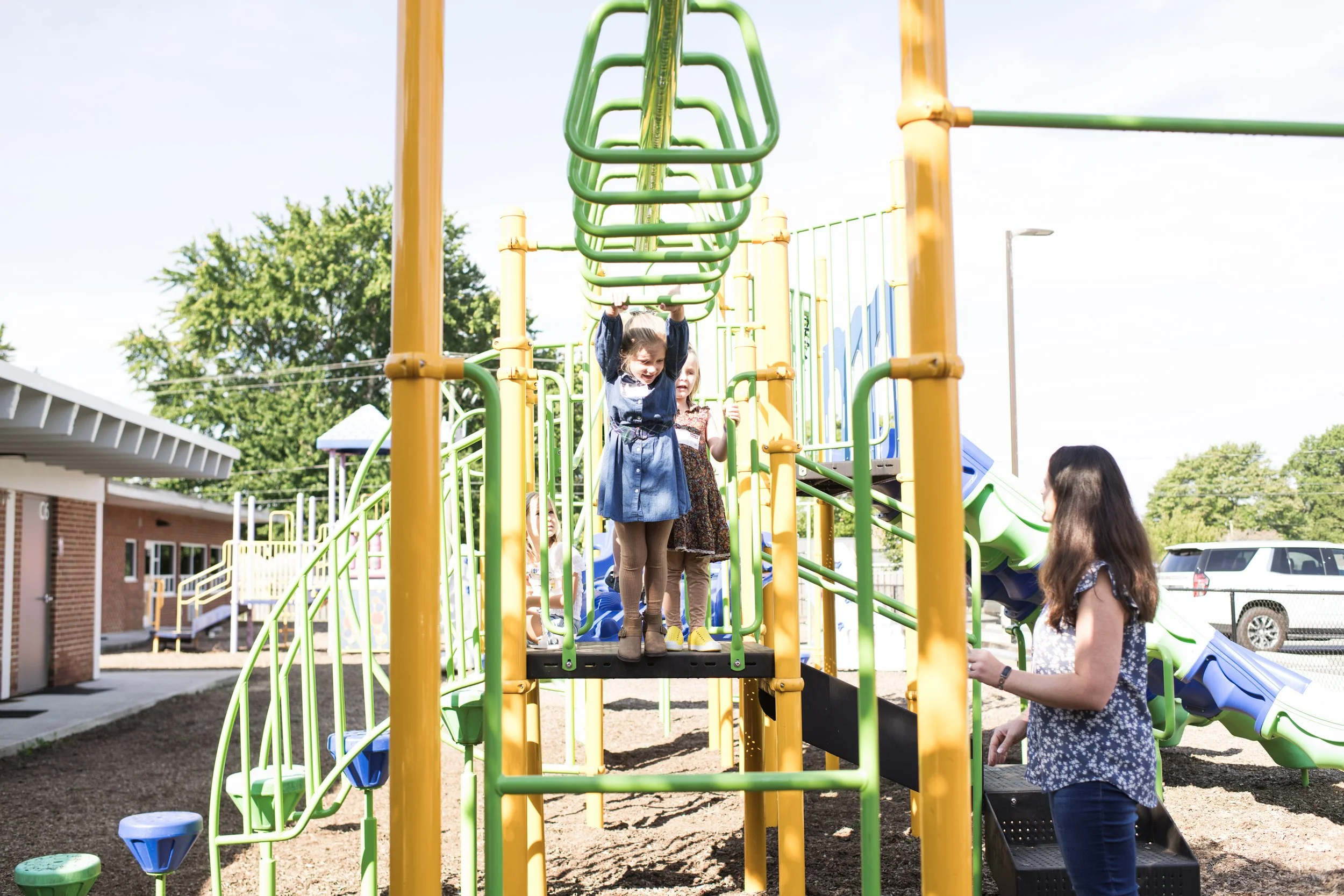 Children playing on a colorful playground with a woman supervising, some children hanging from green bars, a slide to the right, and building in the background on a sunny day.