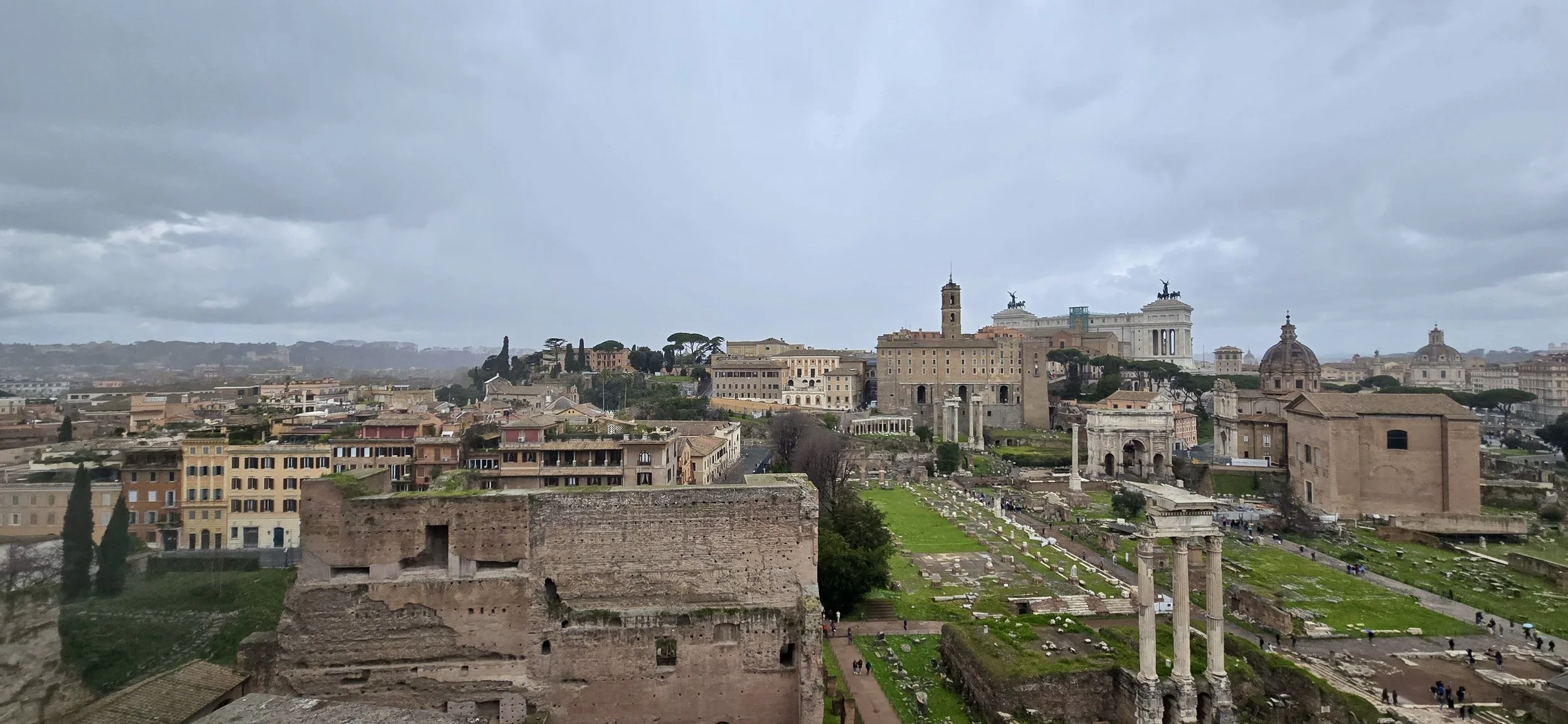 A panoramic view of ancient ruins and historic buildings in Rome, Italy, under a cloudy sky. Visible are ruins, columns, and structures from the Roman Empire, with modern buildings in the background.
