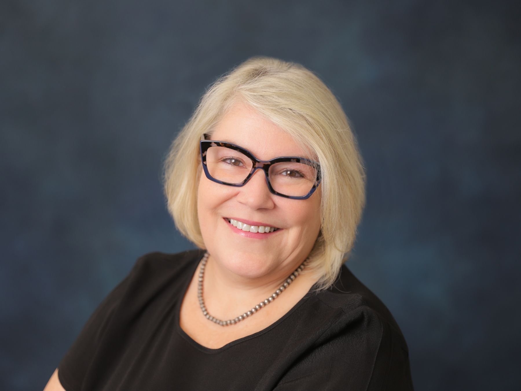 Portrait of a middle-aged woman with blonde hair, glasses, and a black shirt, smiling against a dark blue background.
