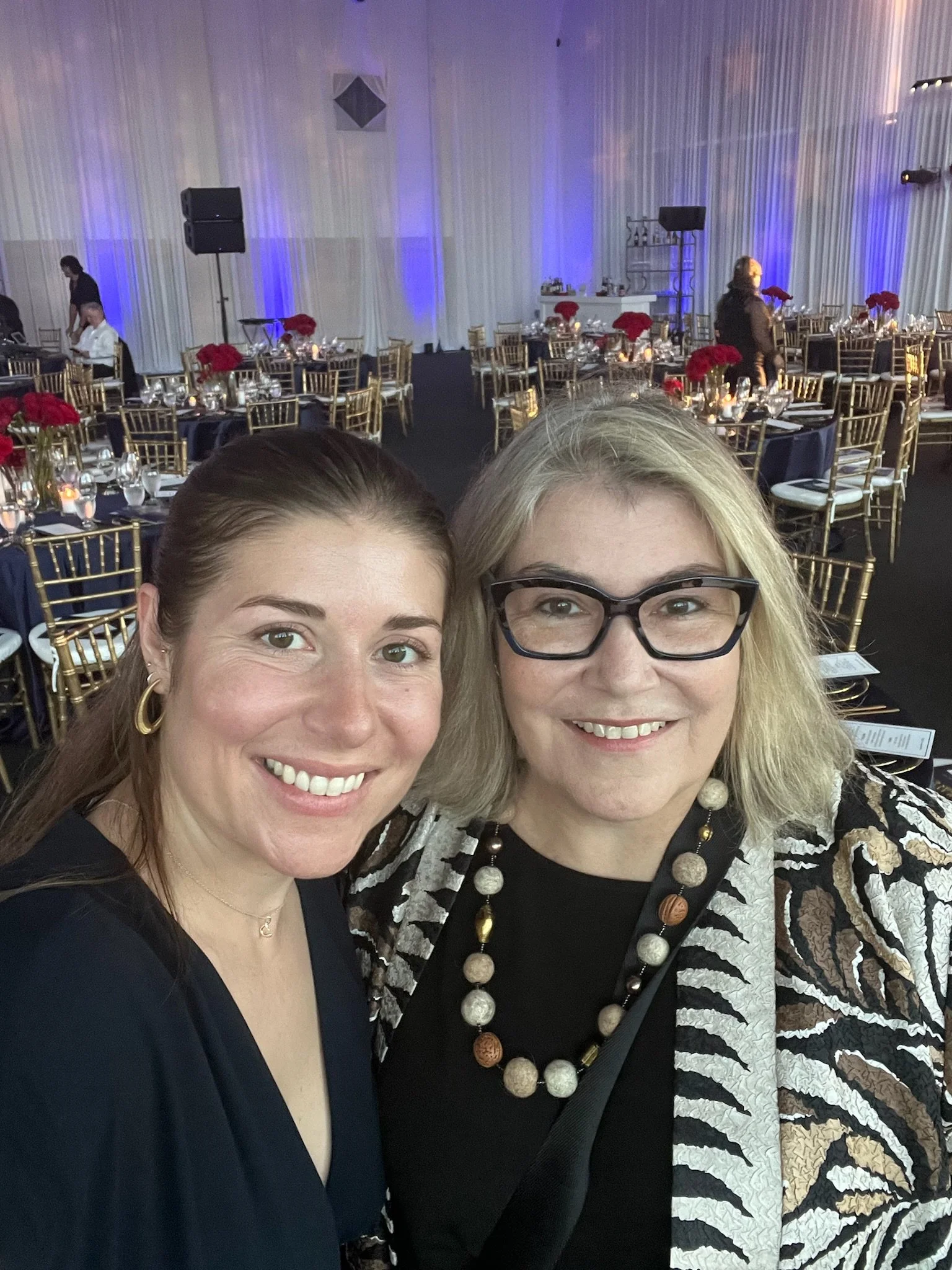 Two women smiling at a formal event with decorated tables and blue lighting in the background.