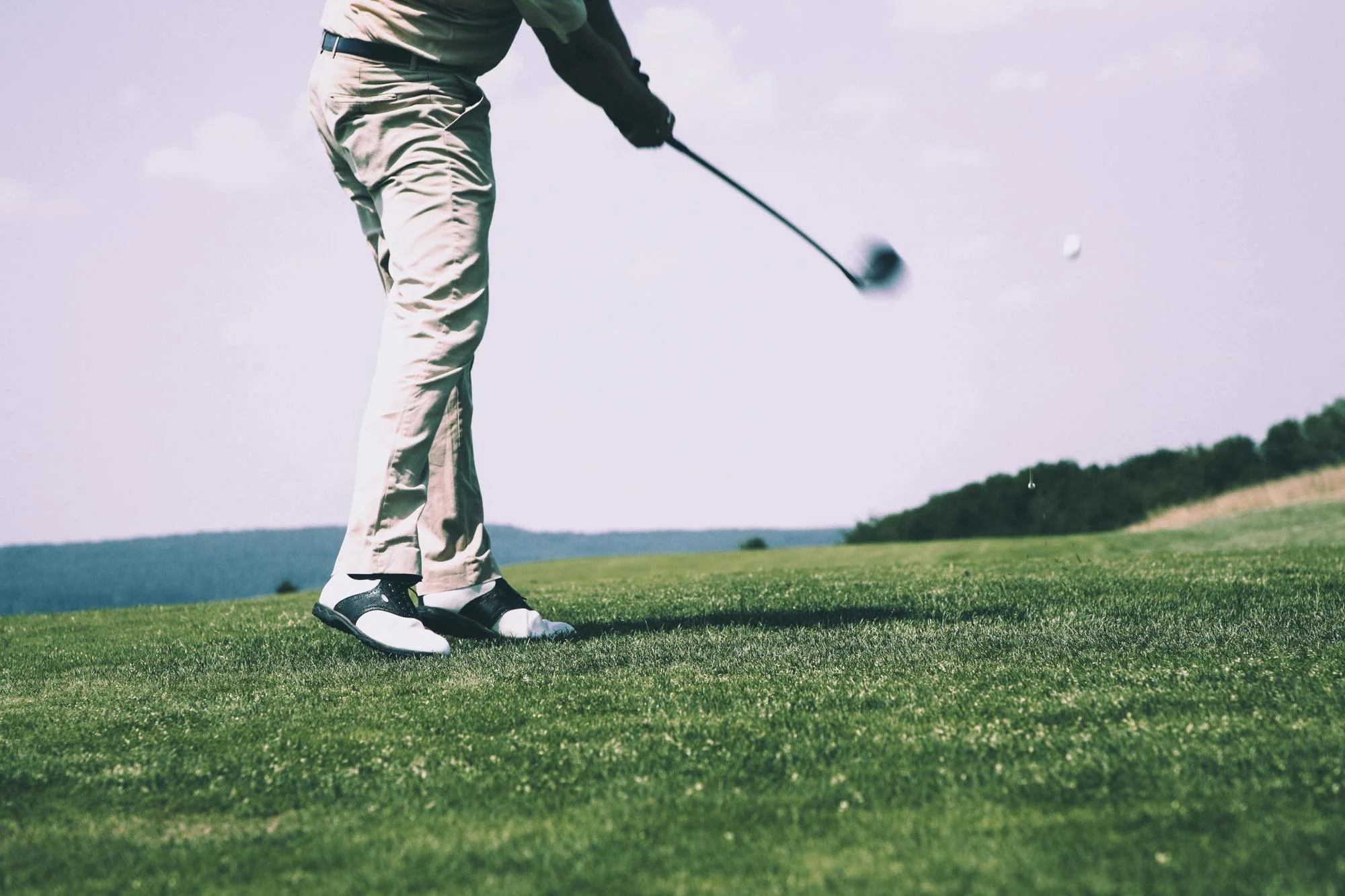 Person in beige pants and black shoes playing golf on a green course with a golf club, preparing to hit the golf ball.