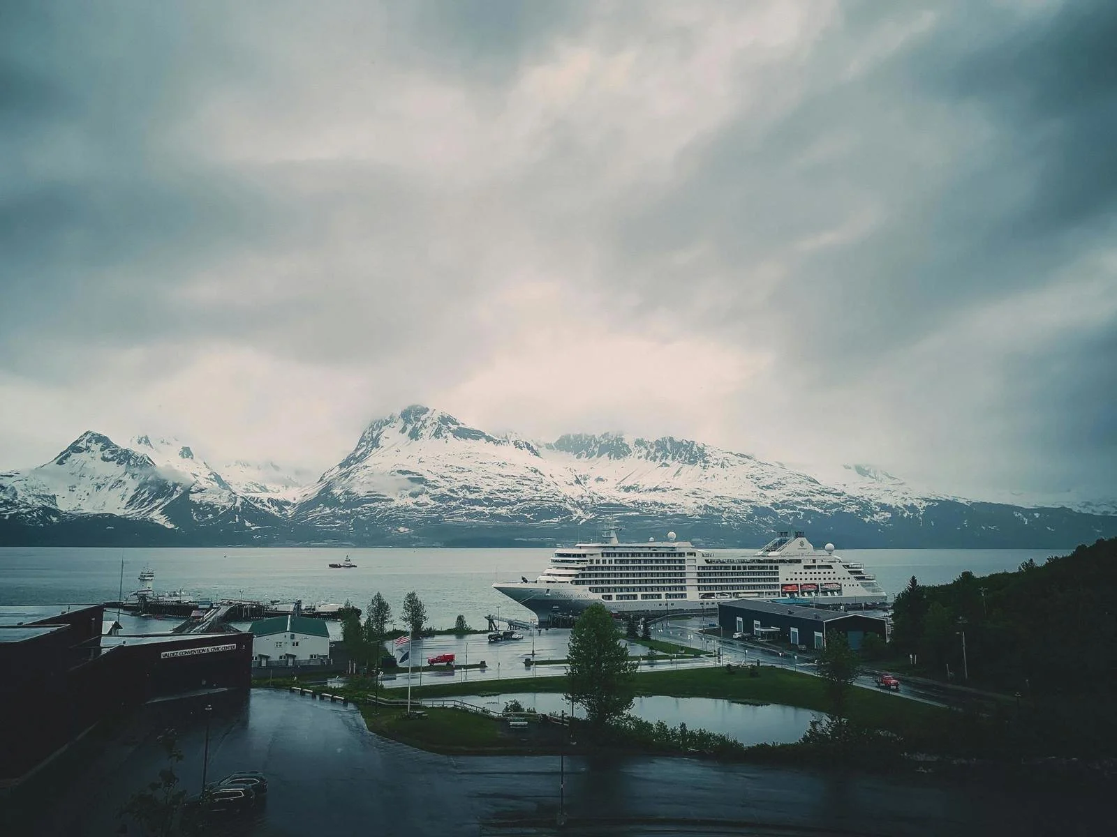 A large cruise ship docked at a harbor with a backdrop of snowy mountains and cloudy sky.