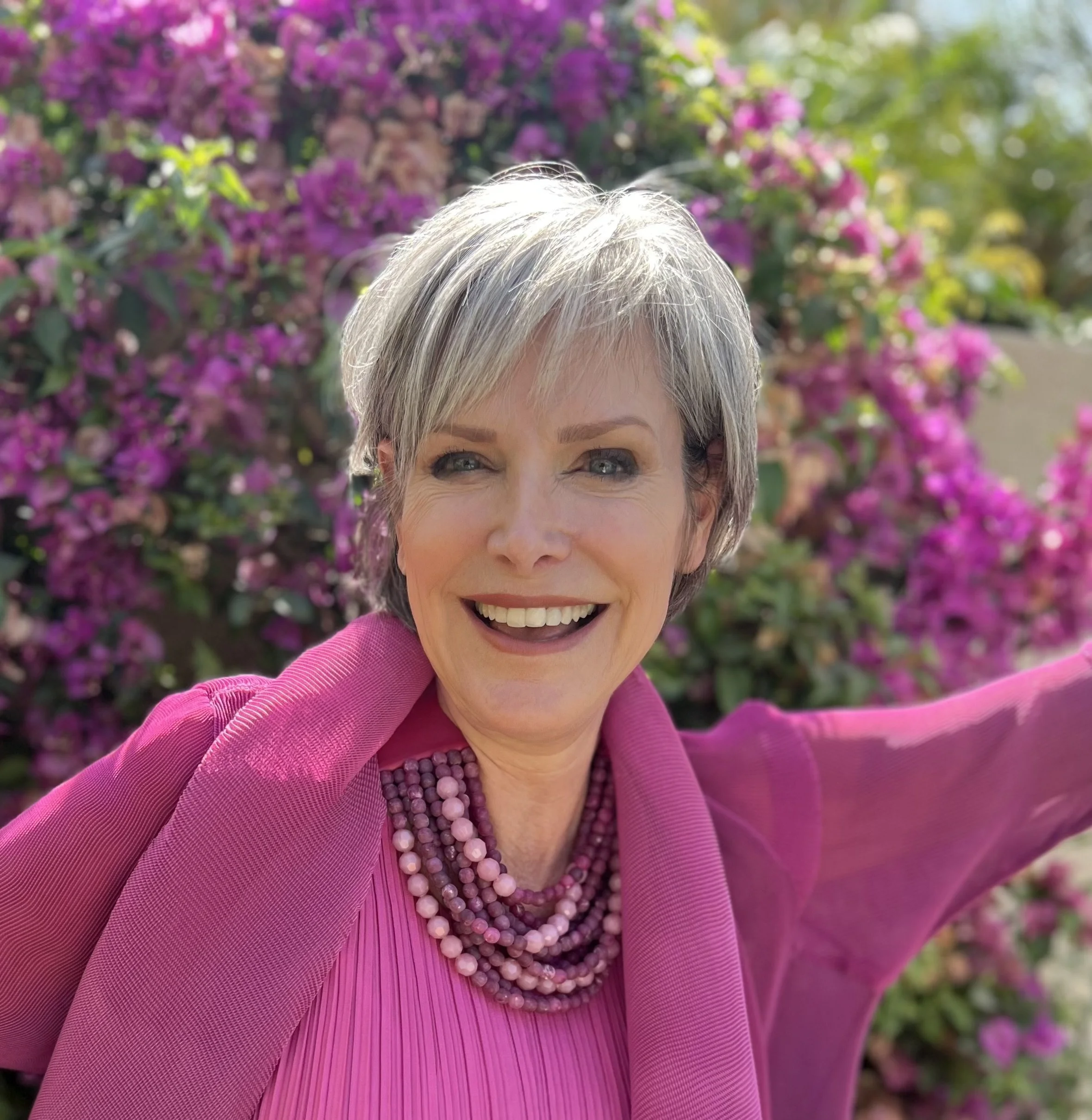 A smiling woman with short gray hair taking a selfie outdoors in front of pink flowering bushes, wearing a pink outfit and beaded necklace.