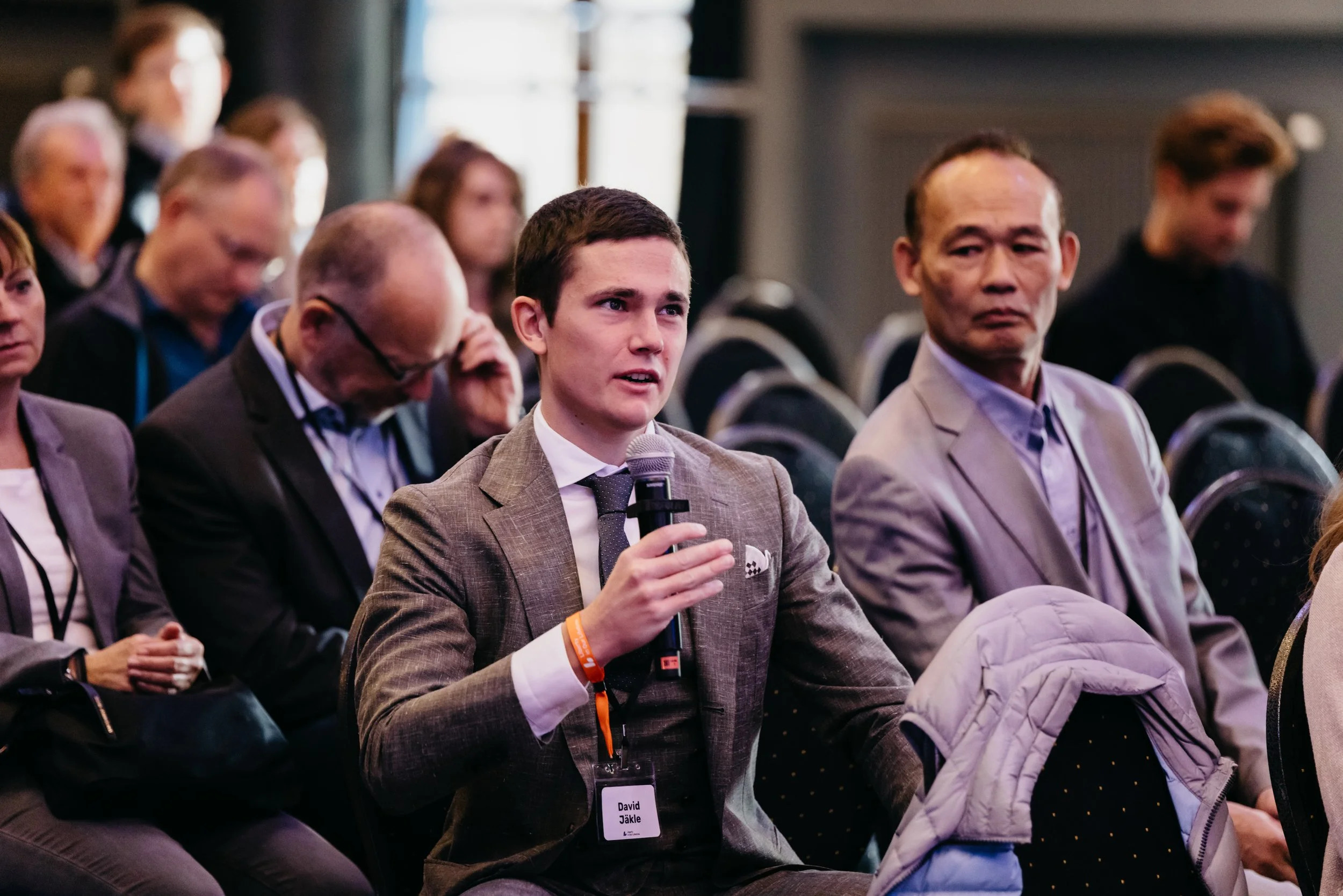 A young man in a gray suit holding a microphone and speaking at a conference. He has a name badge that reads 'David Jäkle.' Other attendees are seated around him, some taking notes, in a dimly lit room with black chairs and a blurred background.