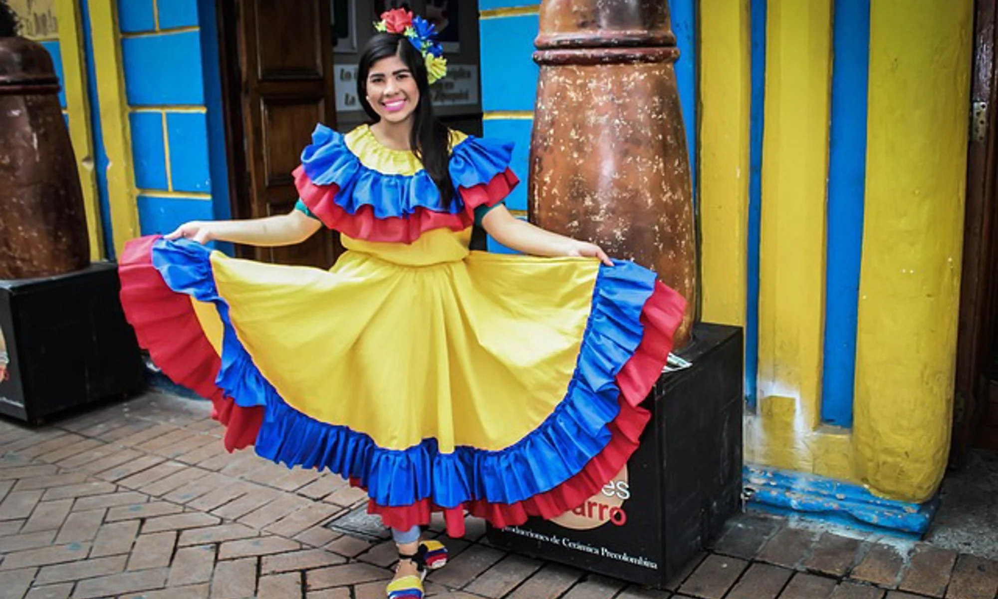 Woman in a colorful traditional dress with yellow, blue, and red ruffles, holding out the skirt to display its full design, standing outdoors on brick pavement in front of brightly painted yellow and blue walls.