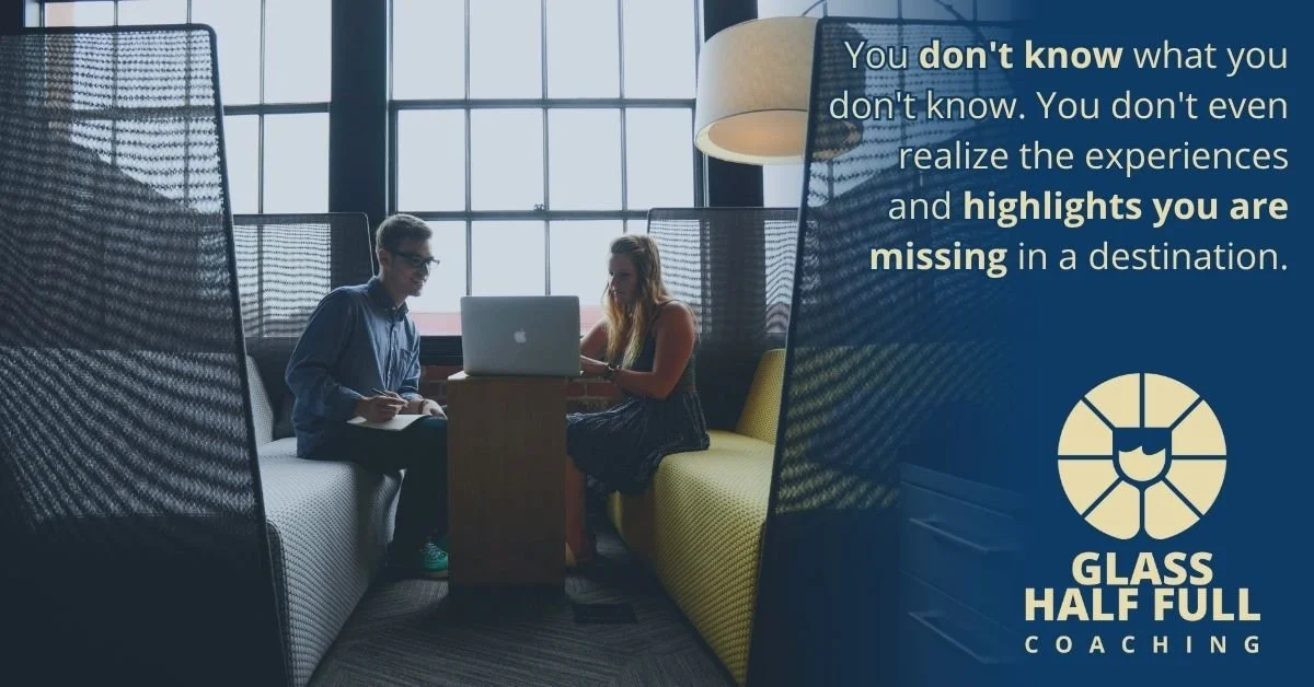 Two people sitting at a table, working on a laptop and taking notes, inside a room with large windows, with a motivational quote on a blue overlay that emphasizes the importance of recognizing missed experiences while traveling, along with a logo for Glass Half Full Coaching.