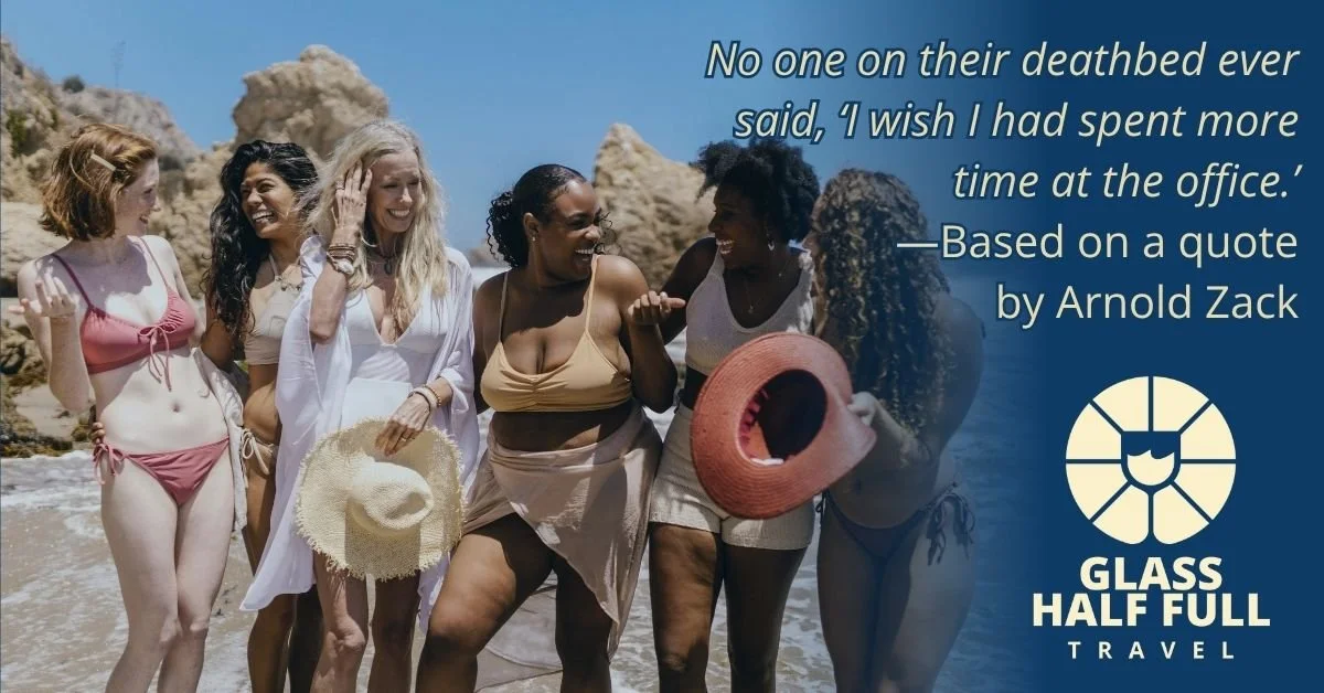 A group of six women in swimsuits and summer clothing standing on the beach, smiling and talking. One woman is holding a straw hat. The background shows rocks and the ocean under a blue sky. The image includes a quote about the office and a logo for Glass Half Full Travel.