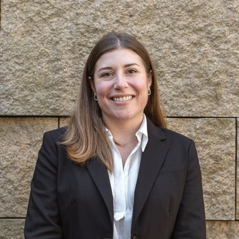 A woman with long brown hair wearing a black blazer and white shirt, posing in front of a stone wall, smiling at the camera.