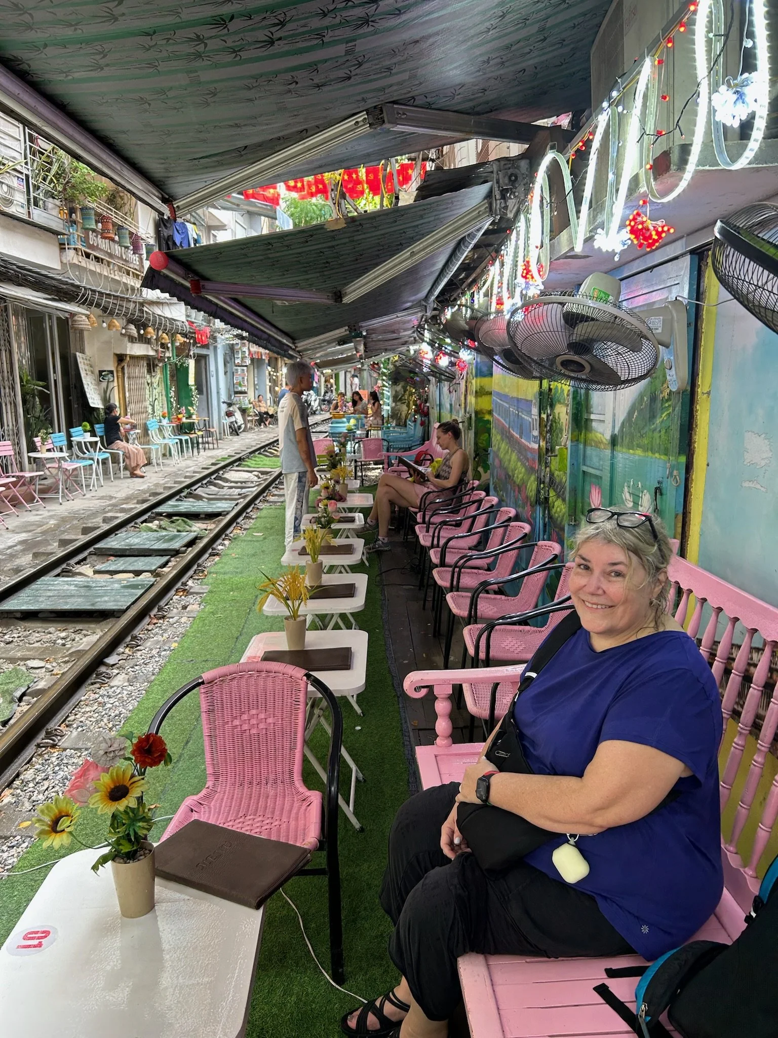 Outdoor railway cafe with pink benches and small tables with flower arrangements, patrons sitting and waiting, colorful street murals, overhead awnings, and decorative lights.