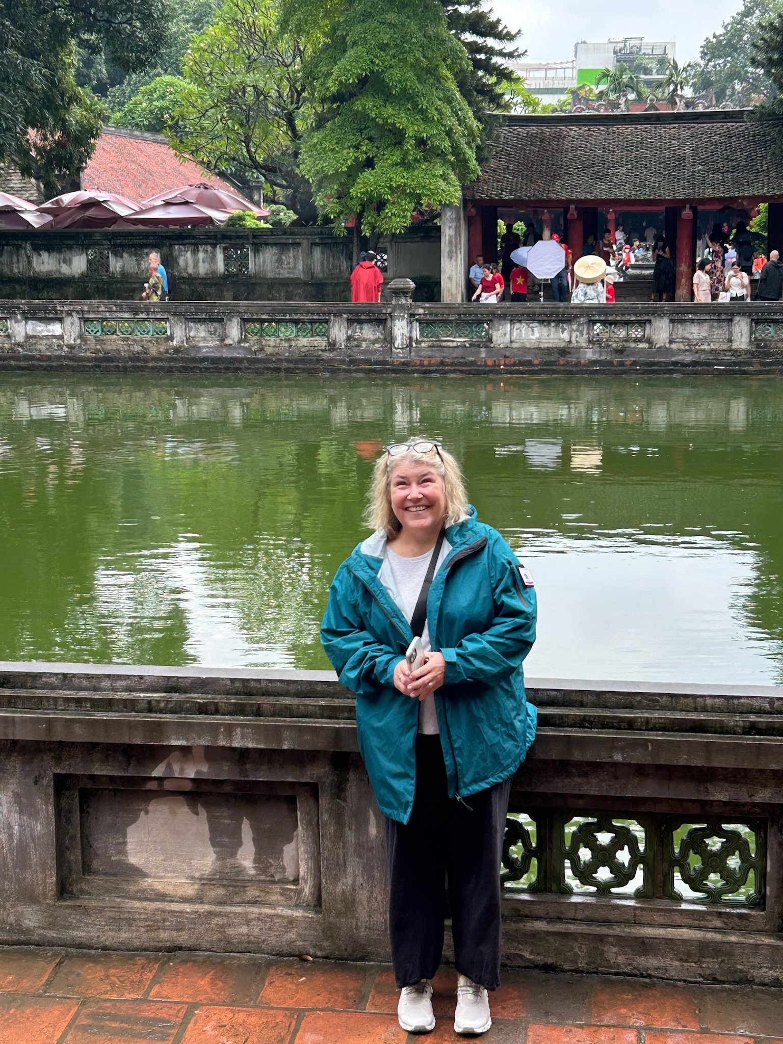 A woman in a blue jacket smiling and standing by a pond with a traditional Asian structure and other people in the background.
